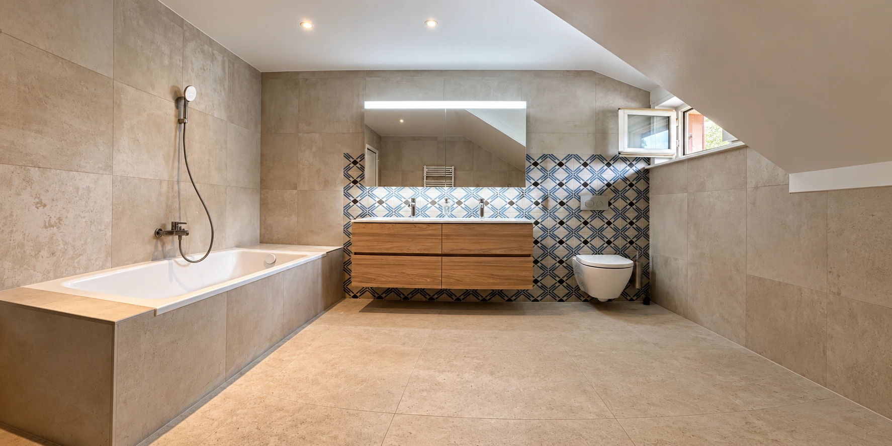 Salle de bains sous les combles : baignoire encastrée, double vasque sur meuble en bois, WC suspendu et carreaux décoratifs bleus